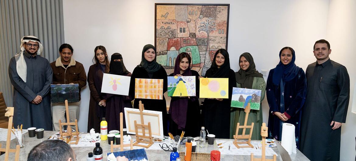 مجموعة من الرجال والنساء، بعضهم يرتدي ملابس الشرق الأوسط التقليدية وأغطية رأس، يقفون خلف طاولة طويلة مليئة بالمواد الفنية، ويعرضون لوحاتهم بفخر. وهم يشاركون في ورشة عمل أو دورة رسم.