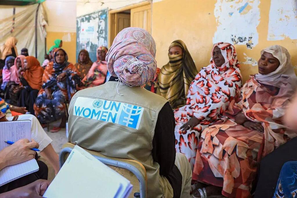 تجلس عضوة في هيئة الأمم المتحدة للمرأة، ترتدي سترة تحمل شعار المنظمة، مع مجموعة من النساء والفتيات السودانيات في اجتماع، لتسلط الضوء على الدعم المقدم للمتضررات من النزاع.