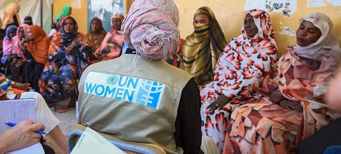 UN Women staff member in a vest with the organization's logo sits with a group of Sudanese women and girls in a meeting, highlighting support for those affected by the conflict.