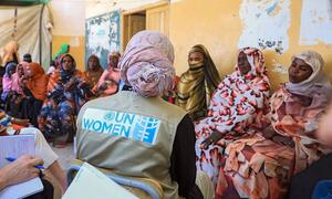 UN Women staff member in a vest with the organization's logo sits with a group of Sudanese women and girls in a meeting, highlighting support for those affected by the conflict.