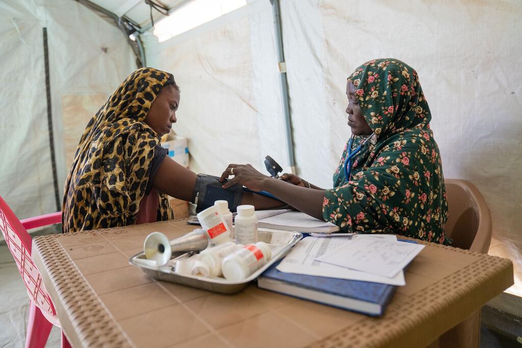 Amina, mulezi aliyehamishwa kutoka Al Fasher, anapima shinikizo la damu ya mwanamke mjamzito katika kituo cha afya kinachoungwa mkono na UNICEF huko Tawila, Darfur Kaskazini, Sudan. Picha inaonyesha Amina akitumia sphygmomanometer kwa mwanamke anayeketi amevaa hijabu yenye muundo wa chui, na vifaa vya matibabu mezani.