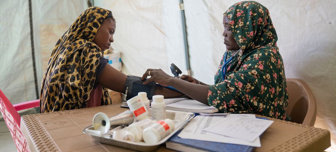 Amina, a midwife displaced from Al Fasher, takes the blood pressure of a pregnant woman at a UNICEF-supported health facility in Tawila, North Darfur, Sudan. The image shows Amina using a sphygmomanometer on a seated woman wearing a leopard print headscarf, with medical supplies on the table.