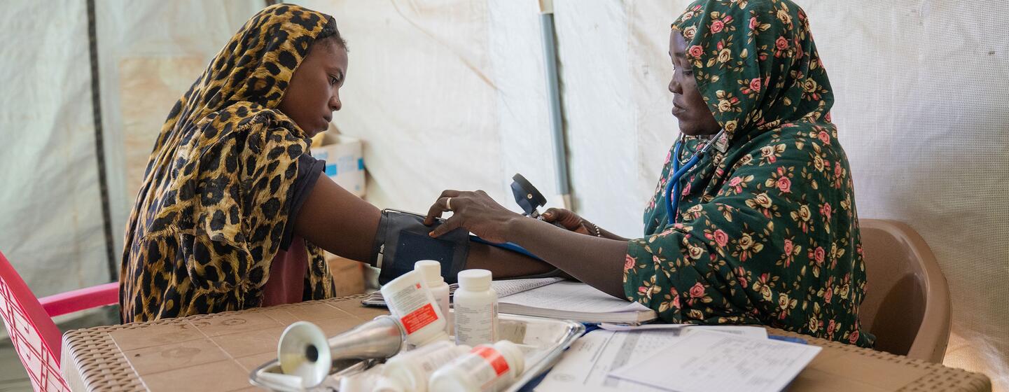 Amina, une sage-femme déplacée d'Al Fasher, mesure la tension artérielle d'une femme enceinte dans un centre de santé soutenu par l'UNICEF à Tawila, dans le Darfour du Nord, au Soudan. L'image montre Amina utilisant un sphygmomanomètre sur une femme assise portant un foulard à motif léopard, avec du matériel médical sur la table.