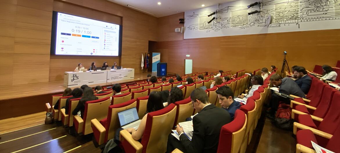 Um público numa sala de conferências num evento modelo da ONU em Lisboa, Portugal, com um painel de oradores na frente e uma apresentação no ecrã.