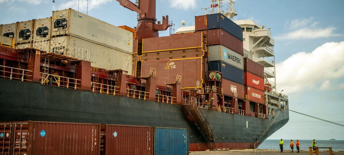 A cargo ship loaded with shipping containers at a port in Tonga. A large crane is visible on the ship's deck, and workers in safety vests are standing on the dock.