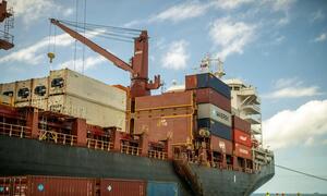A cargo ship loaded with shipping containers at a port in Tonga. A large crane is visible on the ship's deck, and workers in safety vests are standing on the dock.