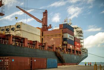 A cargo ship loaded with shipping containers at a port in Tonga. A large crane is visible on the ship's deck, and workers in safety vests are standing on the dock.