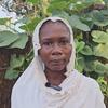 Portrait of a Sudanese woman wearing a white headscarf, standing in front of a thatched wall with green plants.