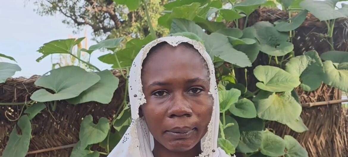 Portrait of a Sudanese woman wearing a white headscarf, standing in front of a thatched wall with green plants.