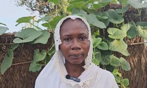 Portrait of a Sudanese woman wearing a white headscarf, standing in front of a thatched wall with green plants.