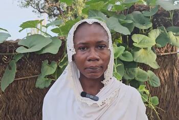 Portrait of a Sudanese woman wearing a white headscarf, standing in front of a thatched wall with green plants.