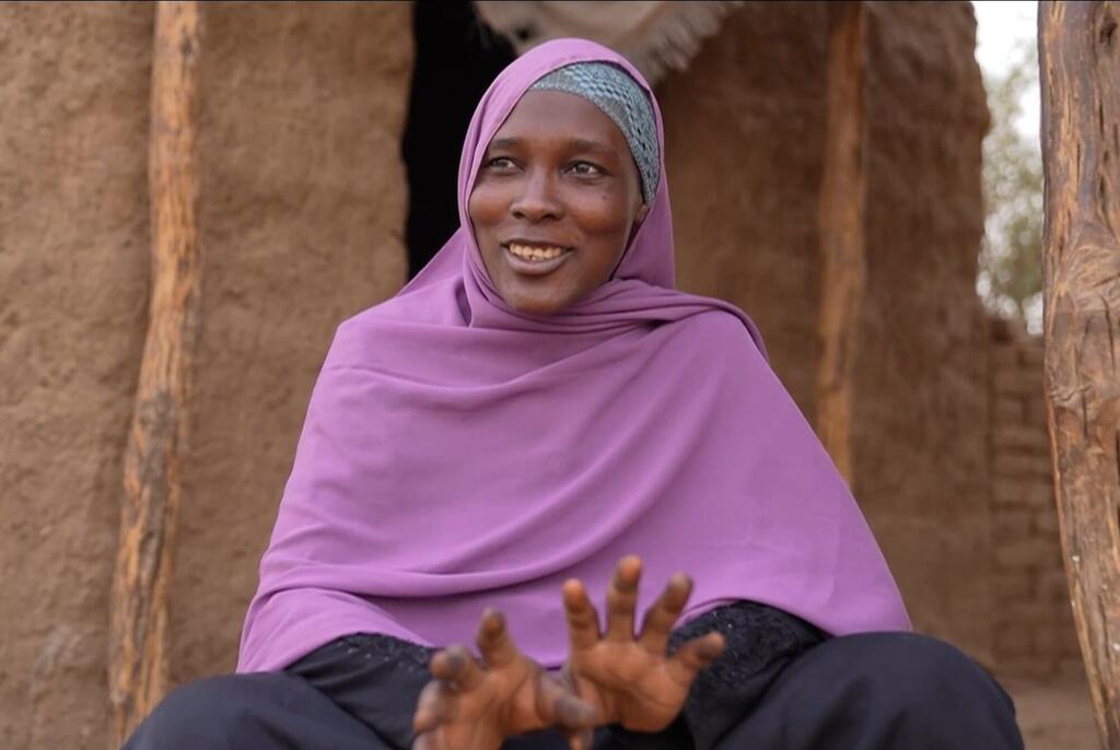 Mwanamke mwenye tabasamu kutoka Sudan akiwa amevaa hijab ya rangi ya zambarau ameketi nje ya nyumba ya udongo, mikono yake ikionyesha ishara wakati akizungumza. Picha hii inahusiana na juhudi za misaada ya kibinadamu nchini Sudan na Chad.