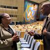 Mwanamume mweusi na mwanamke mweusi wamevaa mavazi rasmi ya biashara wanaozungumza katika ukumbi mkubwa wa mkutano wenye safu za viti tupu. Murali kubwa ya abstrak inaonekana kwenye ukuta katika mandhari ya nyuma.
