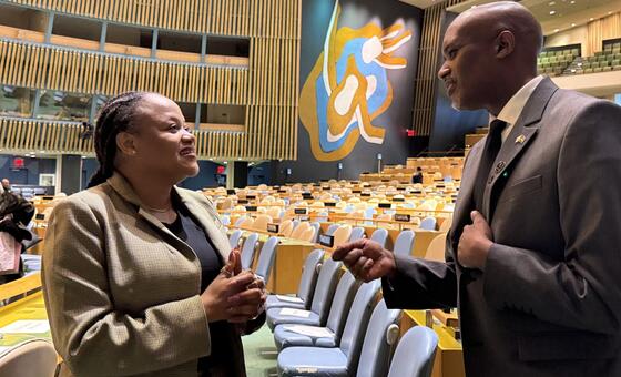 Mwanamume mweusi na mwanamke mweusi wamevaa mavazi rasmi ya biashara wanaozungumza katika ukumbi mkubwa wa mkutano wenye safu za viti tupu. Murali kubwa ya abstrak inaonekana kwenye ukuta katika mandhari ya nyuma.
