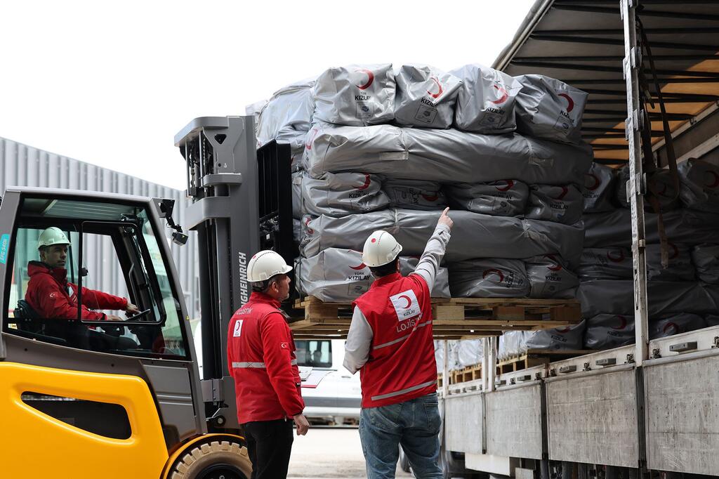 Forkilifu inapakia paleti za vifaa vya misaada ya kibinadamu, ikiwa ni pamoja na hema na blanketi, kwenye lori. Vifaa hivi vimepewa alama ya nembo ya Kizilay (Red Crescent ya Uturuki). Wafanyakazi wa misaada wanaovaa vest nyekundu na kofia za usalama wanamshauri mchakato wa kupakia huko Ankara.