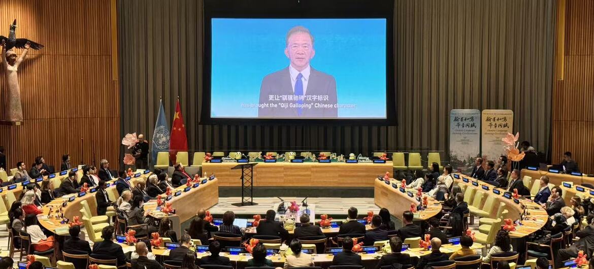 广角镜头展示了一个大型会议厅，厅内代表们坐在弧形桌旁。前方的大屏幕上显示着一位男士正在演讲，屏幕上可见中文文本和英文字幕。房间装饰有独特的木质护墙板和色彩缤纷的吊灯。