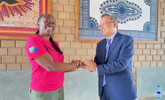 Mwanamke mweusi mwenye tabasamu, amevaa shati la rangi ya waridi, na mwanamume amevaa suti ya bluu, wakishikana mikono mbele ya sanaa ya ukutani ya kitamaduni ya Tanzania.