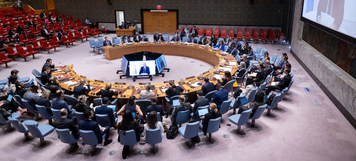 Delegates seated at the UN Security Council table during a meeting on Syria, with Claudio Cordone appearing on screens.