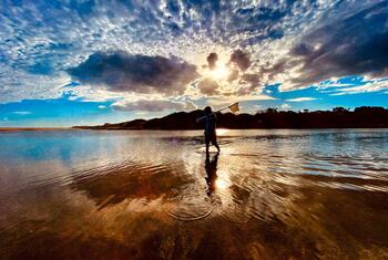 A person stands in shallow water holding a net against a sunset sky in Punta Negra, Maldonado, Uruguay.