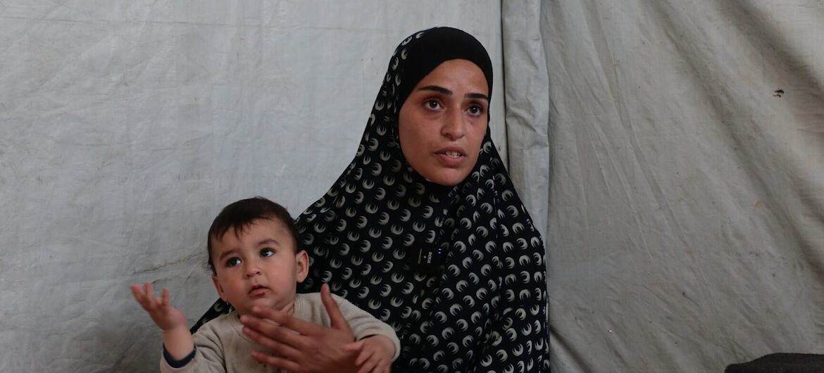 امرأة فلسطينية ترتدي الحجاب تحمل طفلاً داخل خيمة.