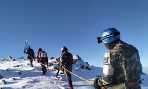 A group of UN peacekeepers in camouflage uniforms and blue helmets conduct a patrol on a snowy mountainous terrain in Syria, holding a UN flag and connected by a safety rope.