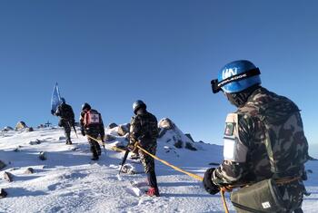 A group of UN peacekeepers in camouflage uniforms and blue helmets conduct a patrol on a snowy mountainous terrain in Syria, holding a UN flag and connected by a safety rope.