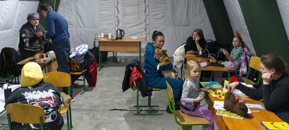 Families with children gather inside a heated mobile 'Point of Invincibility' tent in Kyiv, Ukraine, during a winter power outage. Parents charge devices and prepare hot drinks while children play with toys and modelling clay, supported by UNICEF Psychological First Aid kits and psychosocial services.