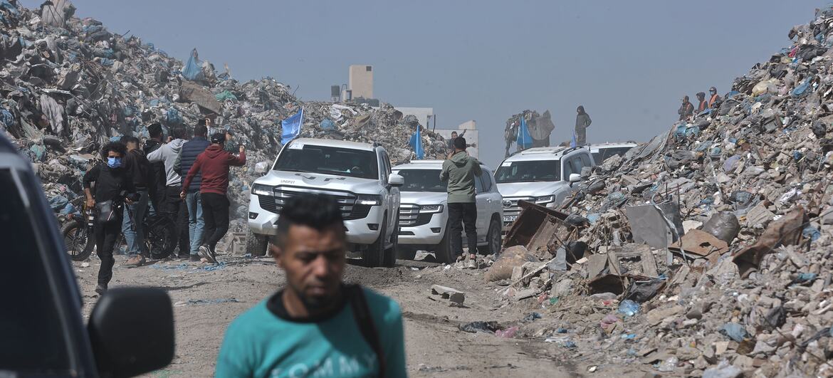 A man walks past a road lined with massive piles of rubble and debris in Gaza, with several white SUVs and people in the background, highlighting the destruction and ongoing relief efforts.