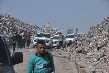 Um homem passa por uma estrada alinhada com enormes pilhas de escombros e detritos em Gaza, com vários SUVs brancos e pessoas no fundo, destacando a destruição e os esforços de socorro em andamento.