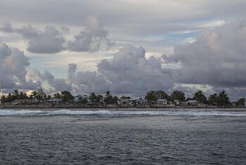 A coastal village on Fale Island in Fakaofo Atoll, Tokelau, with houses and palm trees under a cloudy sky, surrounded by ocean waves.