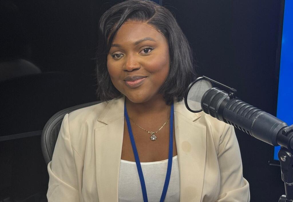 Mwanamke Mwafrika mwenye tabasamu mwenye nywele nyeusi fupi, amevaa blazer ya rangi ya beige na lanyard ya bluu, akizungumza kwenye kipaza sauti cha studio ya kitaalamu.