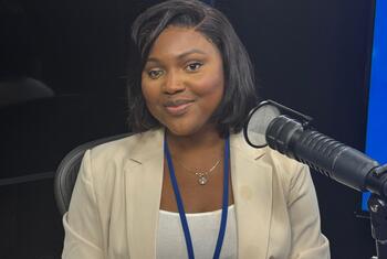 Mwanamke Mwafrika mwenye tabasamu mwenye nywele nyeusi fupi, amevaa blazer ya rangi ya beige na lanyard ya bluu, akizungumza kwenye kipaza sauti cha studio ya kitaalamu.