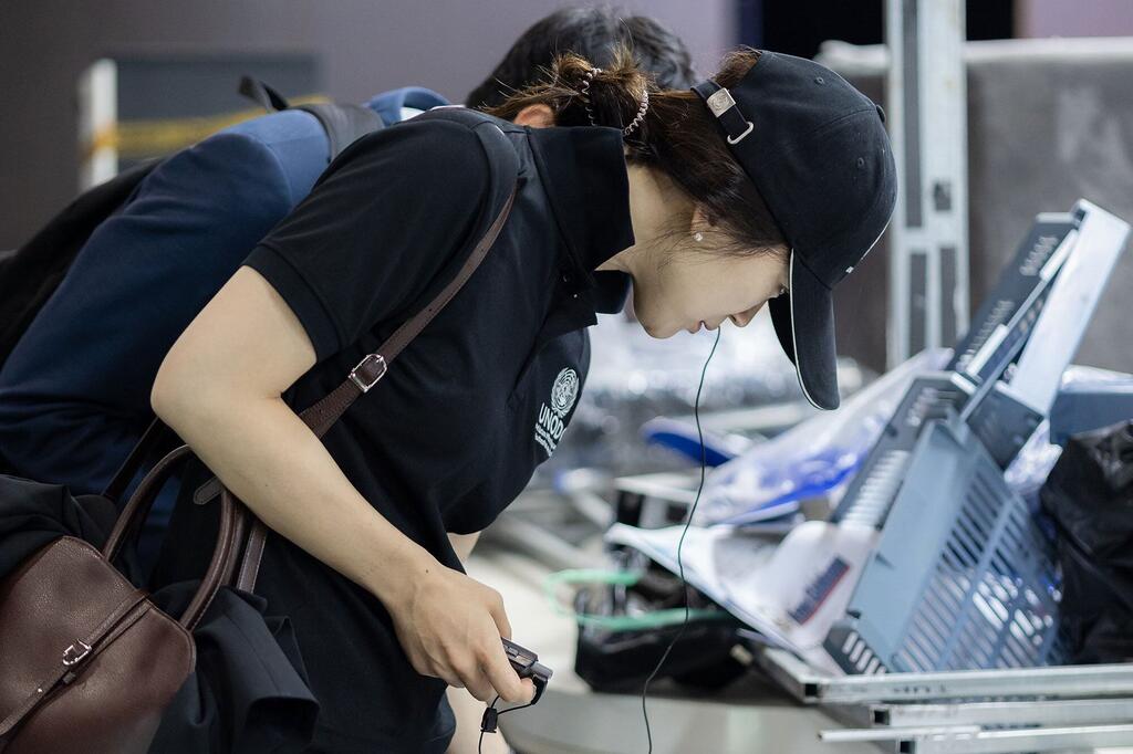 Une femme de sécurité aux traits asiatiques, vêtue d'une casquette noire et d'un polo avec un logo de l'ONU, se concentre sur une tâche de contrôle des bagages à l'aéroport.