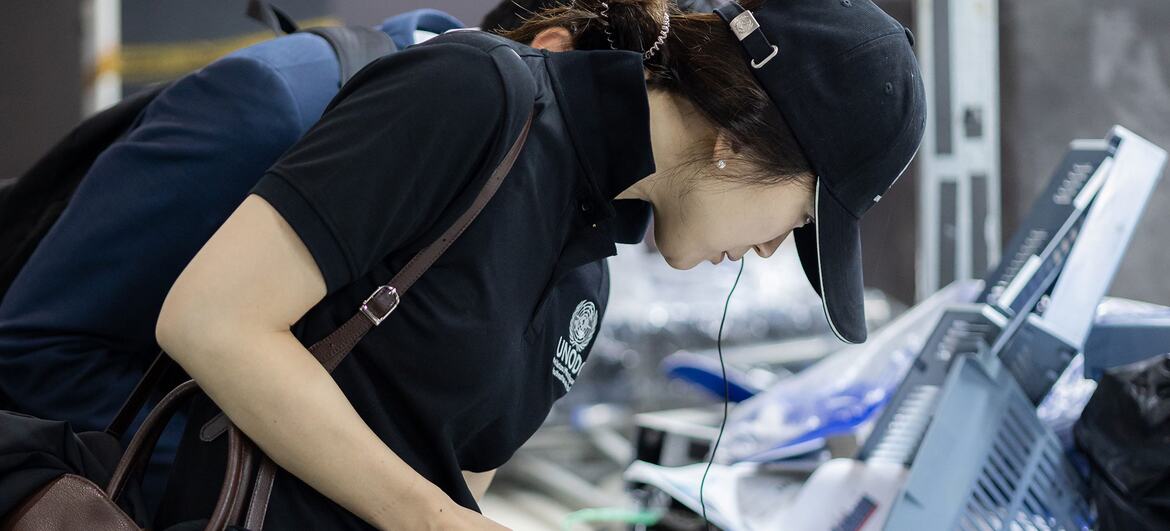 A female security officer with Asian features, wearing a black cap and polo shirt with a UN logo, is focused on a baggage screening task at an airport.