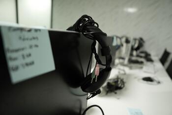 A close-up view of a pair of black wired headphones with pink and green audio jacks hanging from the side of a black computer monitor in an office or computer lab setting.