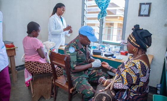Mlinzi wa amani wa Tanzania wa MONUSCO aliyevaa yunifomu ya kujificha na kofia ya rangi ya bluu ya Umoja wa Mataifa anatoa huduma ya matibabu kwa mwanamke katika kliniki katika Jamhuri ya Kidemokrasia ya Kongo, wakati mwanafunzi wa afya anayevaa koti la rangi ya nyeupe anatazama uchunguzi huo.