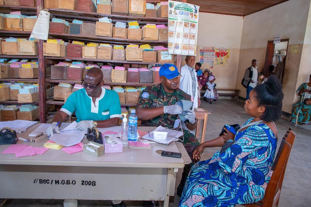 Wanajeshi wa kudumisha amani wa Tanzania kutoka MONUSCO wanasaidia katika usajili wa wagonjwa katika kliniki ya afya nchini DRC. Mwanajeshi wa kudumisha amani mwenye kofia ya UN ya bluu na mtaalamu wa afya wameketi kwenye dawati pamoja na mgonjwa wa kike, wakiwa wamezungukwa na rafu za rekodi za matibabu.