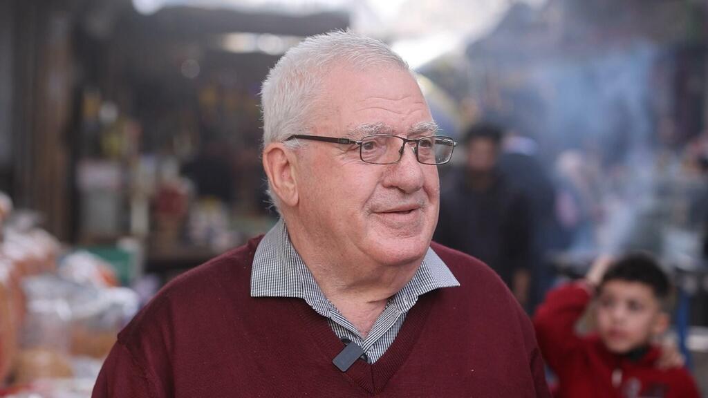 يُجري مقابلة مع رجل مسن يرتدي النظارات والسترة الحمراء في سوق مزدحمة في غزة خلال شهر رمضان.