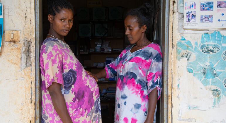 मितीके डेस्टा, एक गर्भवती महिला, एक दरवाजे पर खड़ी है, जबकि स्वास्थ्य विस्तार कार्यकर्ता येशीवर्क बेलै ने उसके पेट पर एक आरामदायक हाथ रखा है, जो इथियोपिया में आवश्यक मातृ देखभाल सुनिश्चित करता है।