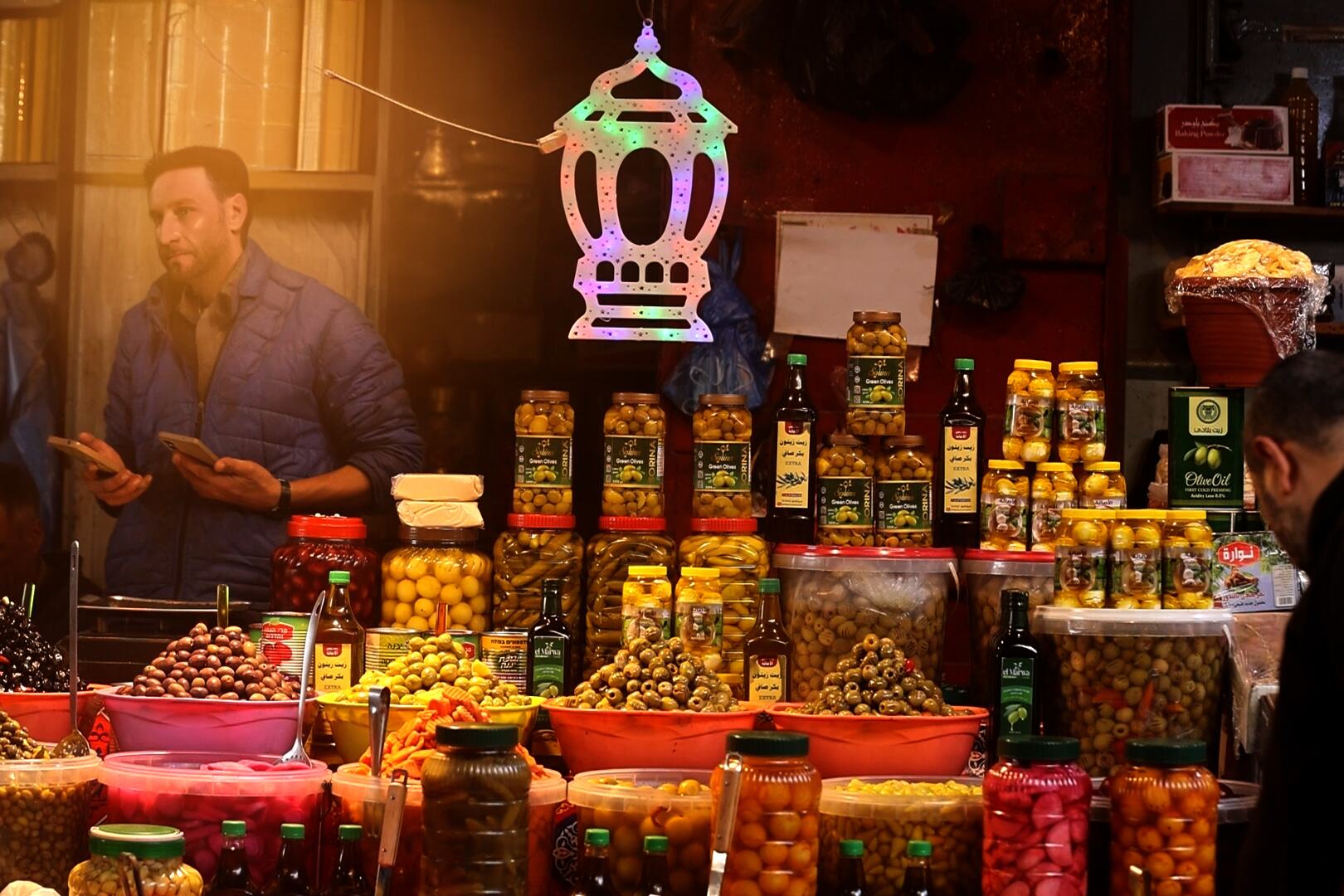 غزہ میں ایک مصروف رمضان منڈی میں مٹھائیوں، کھجوروں اور ضروری غذائی اشیا کی ایک وسیع رینج پیش کی گئی ہے، اور اسٹالوں کے اوپر ایک رنگا رنگ لالٹین لٹکی ہوئی ہے۔