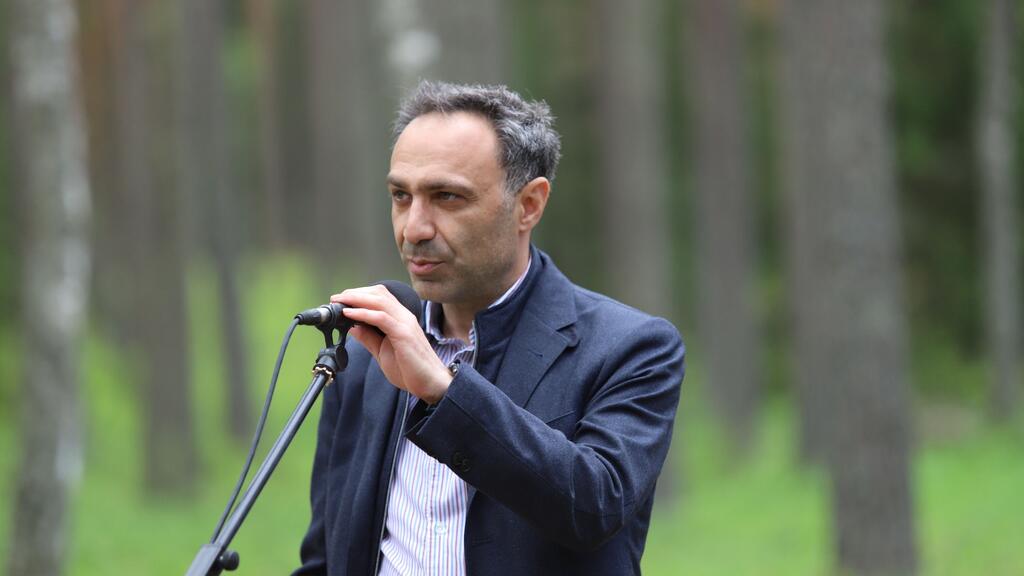 A man in a suit speaking into a microphone in a forest setting.