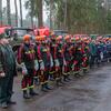 Una fila de bomberos bielorrusos con uniforme y cascos naranjas de pie frente a camiones de bomberos en un bosque.