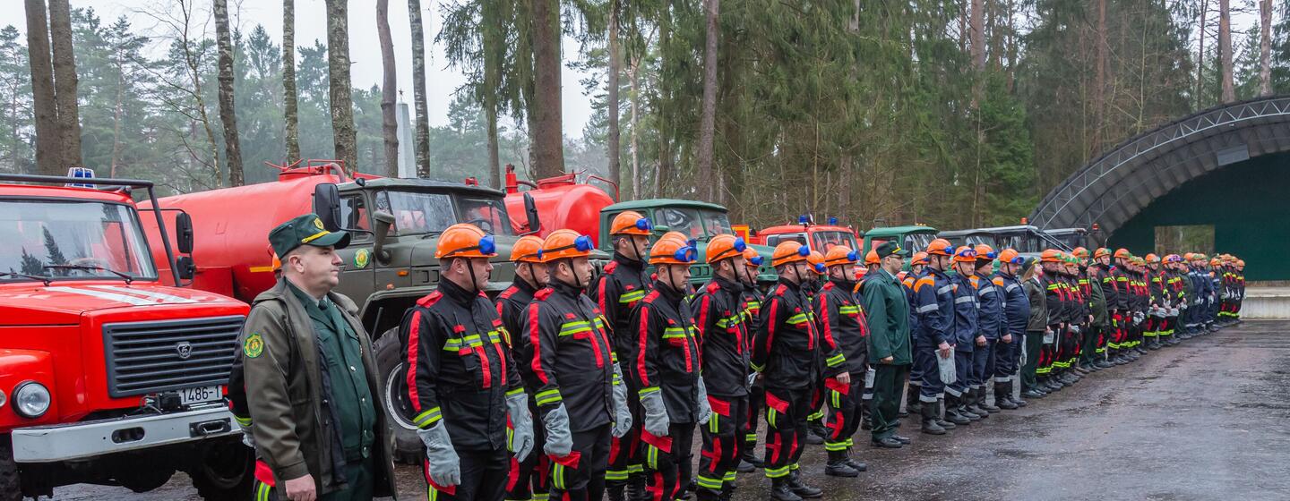 Una fila de bomberos bielorrusos con uniforme y cascos naranjas de pie frente a camiones de bomberos en un bosque.
