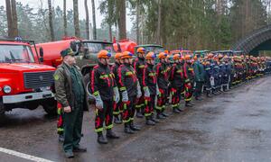 Una fila de bomberos bielorrusos con uniforme y cascos naranjas de pie frente a camiones de bomberos en un bosque.