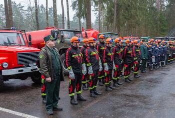 Una fila de bomberos bielorrusos con uniforme y cascos naranjas de pie frente a camiones de bomberos en un bosque.