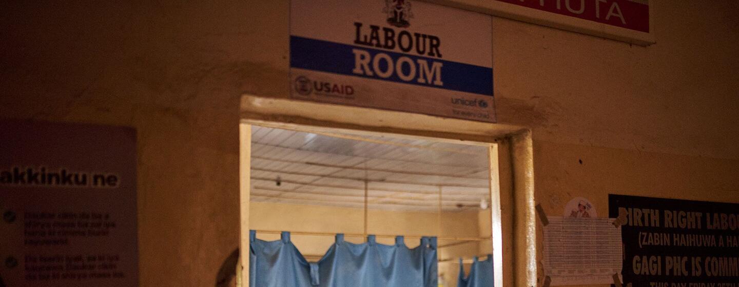 Hauwa Yusuf, 30 ans, tenant son nouveau-né dans la salle de travail du centre de santé primaire de Gagi, dans l'État de Sokoto, au Nigeria. Un panneau au-dessus de la porte indique " LABOUR ROOM " et montre les logos de l'USAID et de l'UNICEF.