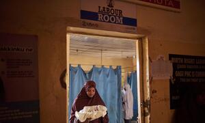 Hauwa Yusuf, de 30 años, sostiene a su bebé recién nacido dentro de la sala de parto del Centro de Salud Primaria Gagi en el estado de Sokoto, Nigeria. Un letrero encima de la puerta dice 'LABOUR ROOM' y muestra logotipos de USAID y UNICEF.