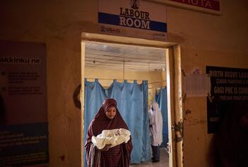 Hauwa Yusuf, de 30 años, sostiene a su bebé recién nacido dentro de la sala de parto del Centro de Salud Primaria Gagi en el estado de Sokoto, Nigeria. Un letrero encima de la puerta dice 'LABOUR ROOM' y muestra logotipos de USAID y UNICEF.