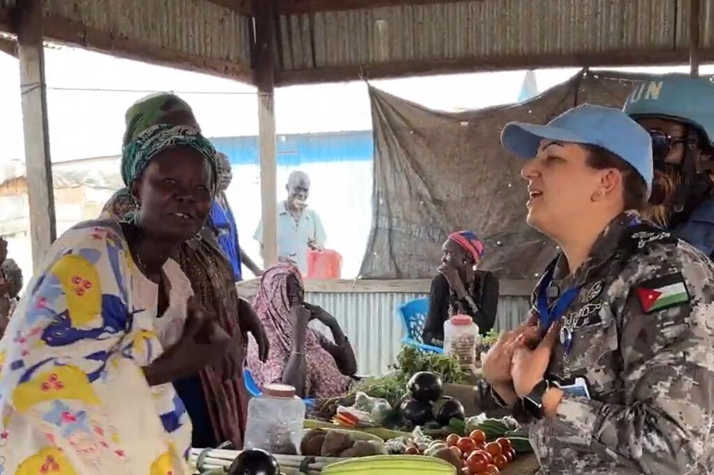 Mwanamke mmoja katika kikosi cha kulinda amani cha Umoja wa Mataifa akiwa amevaa yunifomu ya Jordan na kofia ya chuma ya bluu akizungumza na mwanamke mmoja kutoka Sudan Kusini katika kibanda cha soko kilichojaa mboga safi.