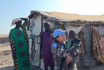 Mwanamke mmoja katika kikosi cha kulinda amani cha Umoja wa Mataifa akiwa amevaa yunifomu ya kujificha na kofia ya chuma ya bluu akichukua mtoto mwenye tabasamu huku amesimama mbele ya makao ya muda pamoja na wanawake wawili wa eneo hilo huko Bentiu, Sudan Kusini.